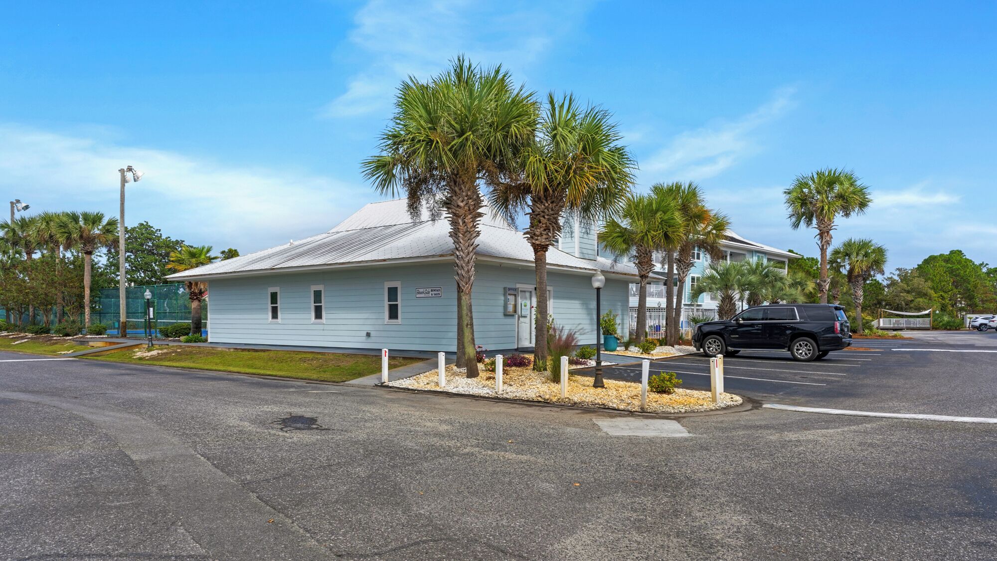 A light blue building with a white roof, palm trees, and a black SUV parked in front.