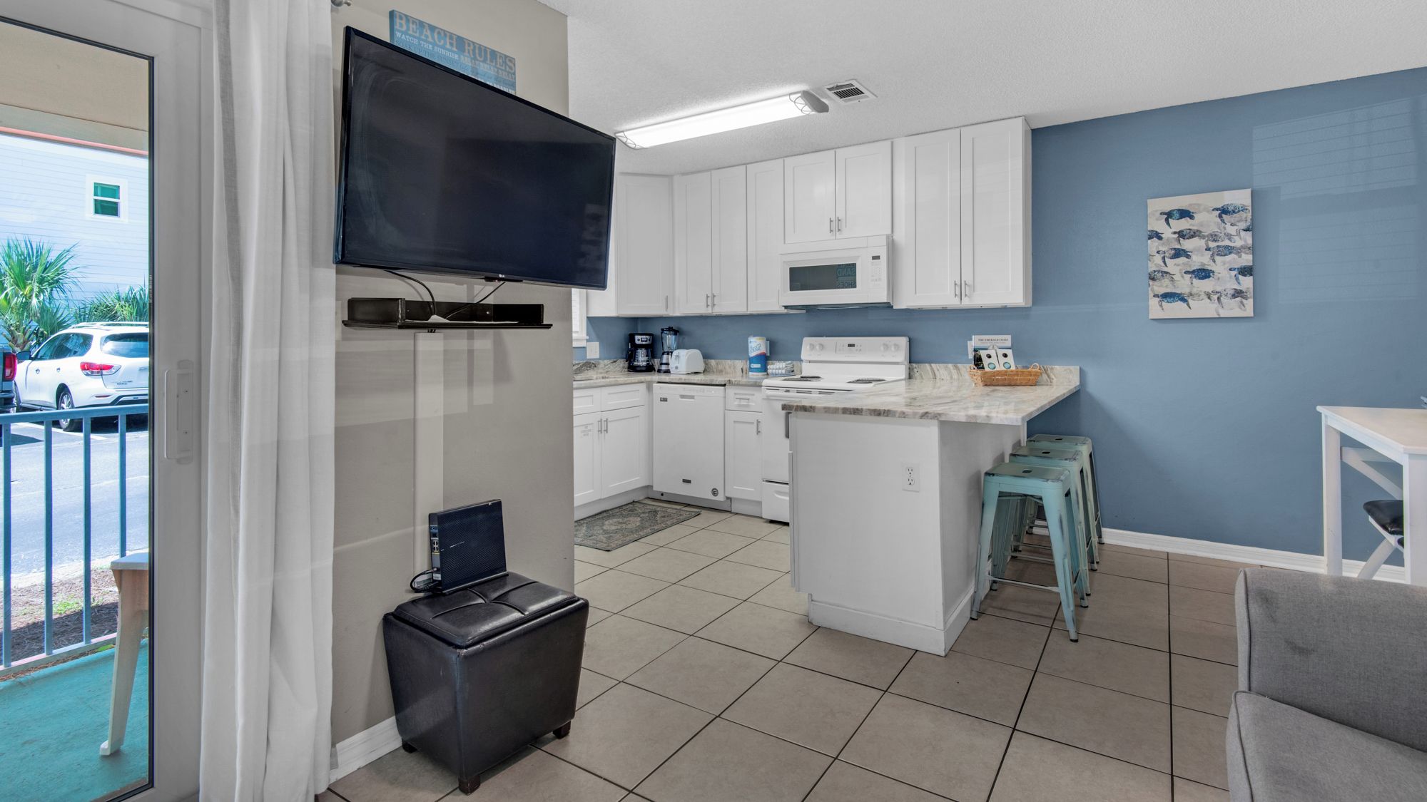 A compact kitchen with white cabinets, a microwave, and a small island. There is a television mounted above the counter and a painting on the wall.
