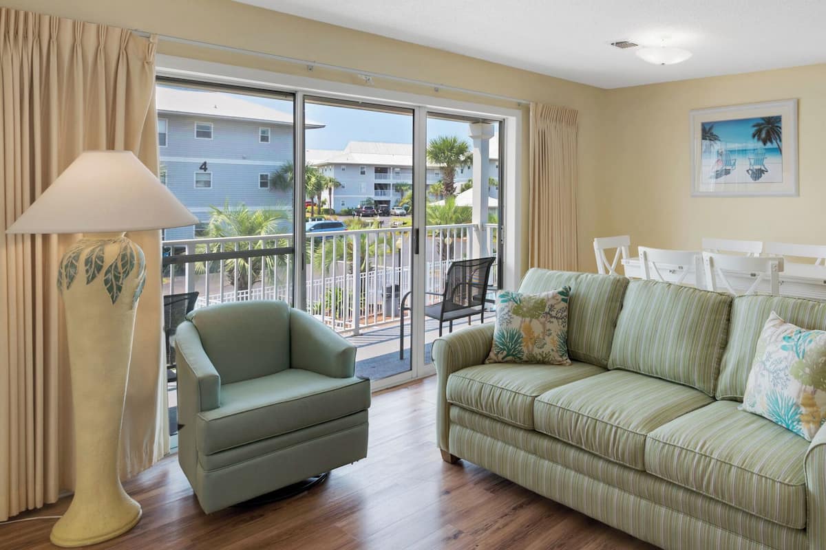 A living room with a green sofa, a chair, a lamp, and a balcony with a view of buildings and palm trees.