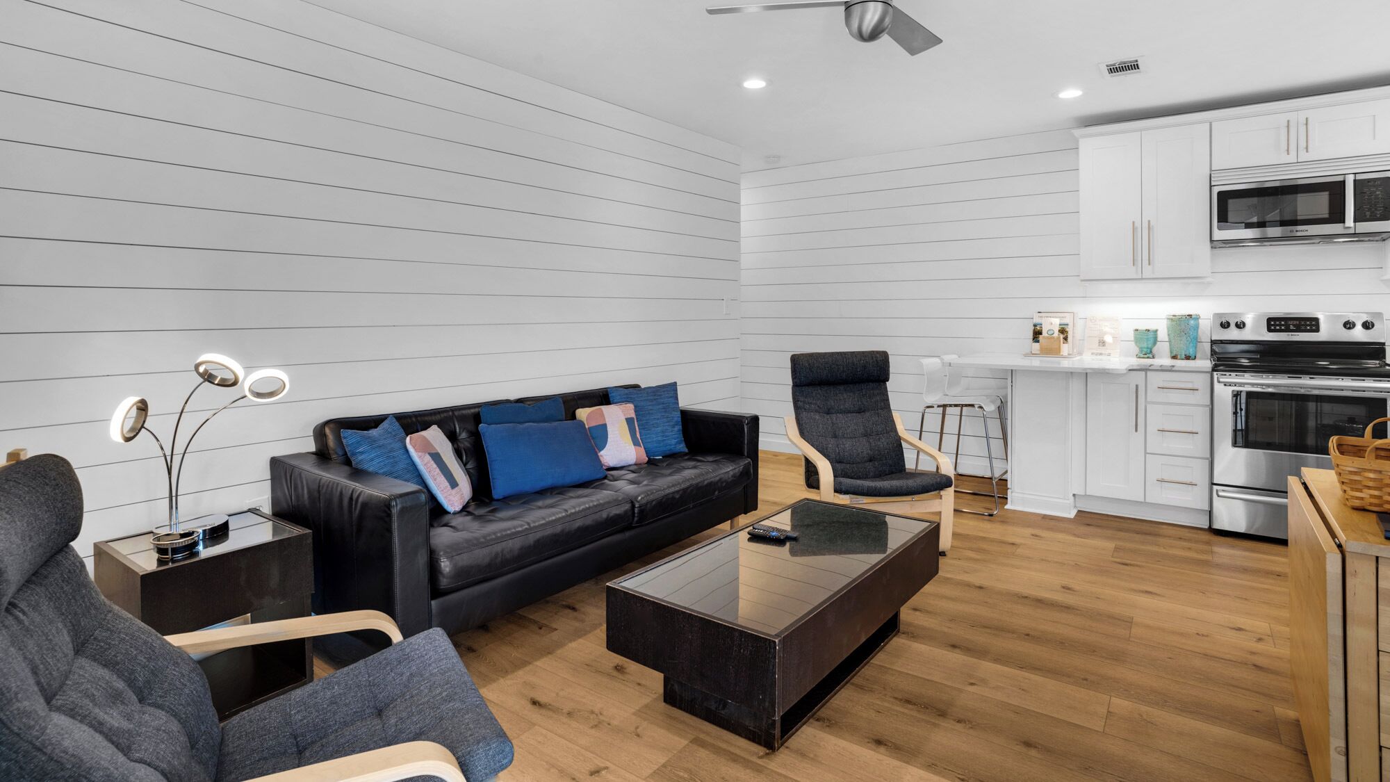 A modern living room with a black leather sofa, grey armchairs, and a glass coffee table. A kitchen with white cabinets and stainless steel appliances is visible in the background.