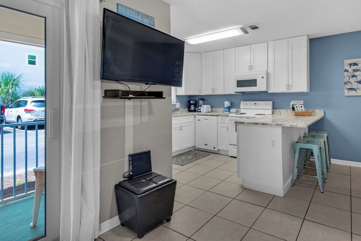 A compact kitchen with white cabinets, a microwave, and a small island. There is a television mounted above the counter and a painting on the wall.