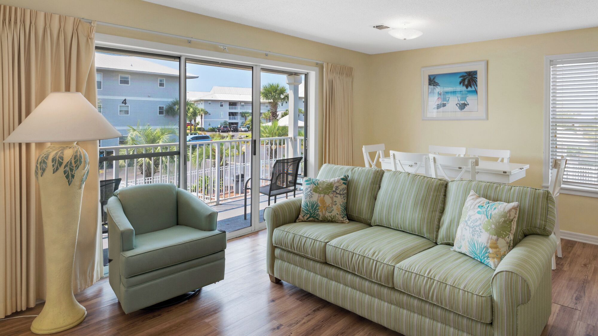 A living room with a green sofa, a chair, a lamp, and a balcony with a view of buildings and palm trees.