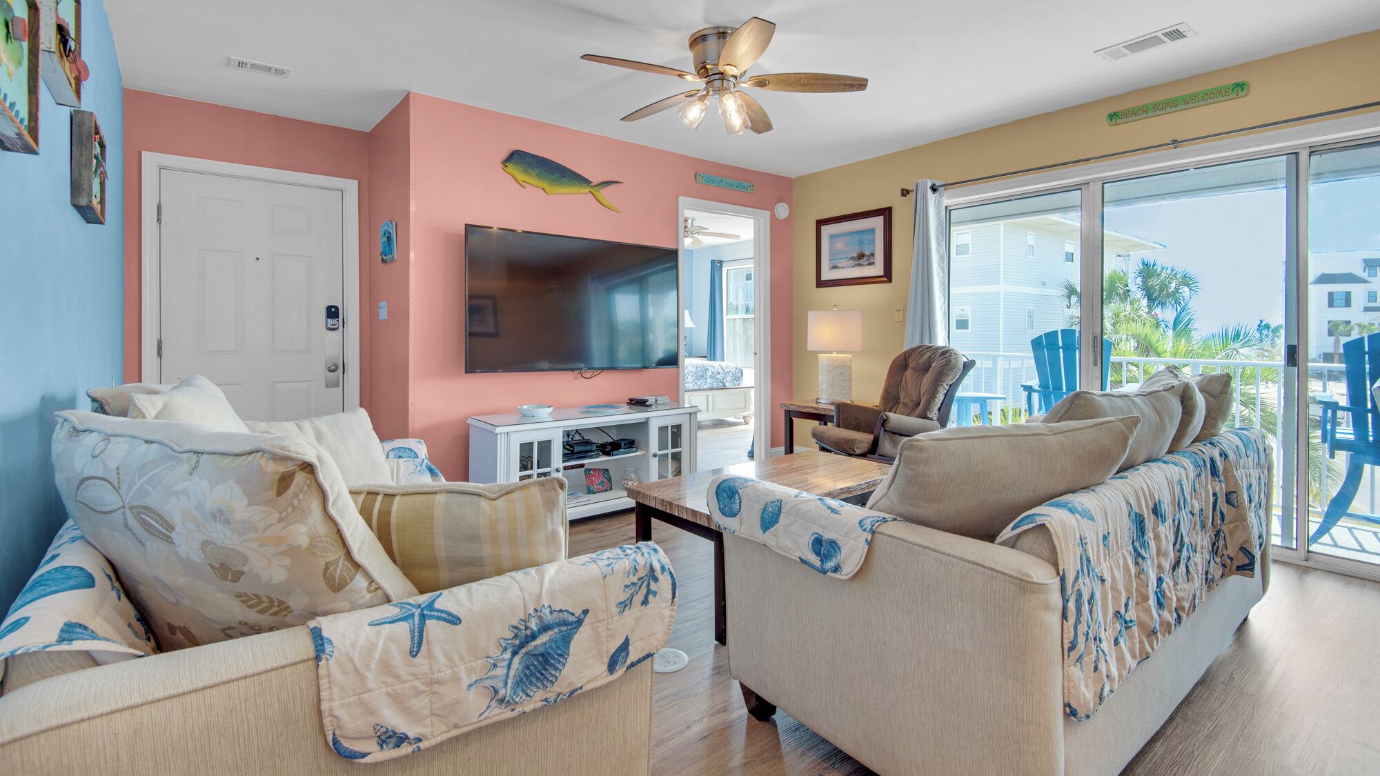 A living room with a TV, sofa, and chairs, a ceiling fan, and a view of the outside.