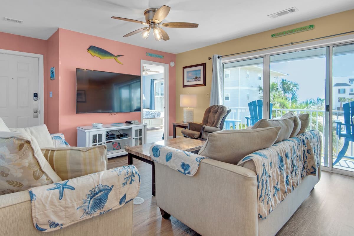 A living room with a TV, sofa, and chairs, a ceiling fan, and a view of the outside.