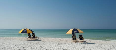Two beach umbrellas with lounge chairs on a sandy beach with clear blue water.