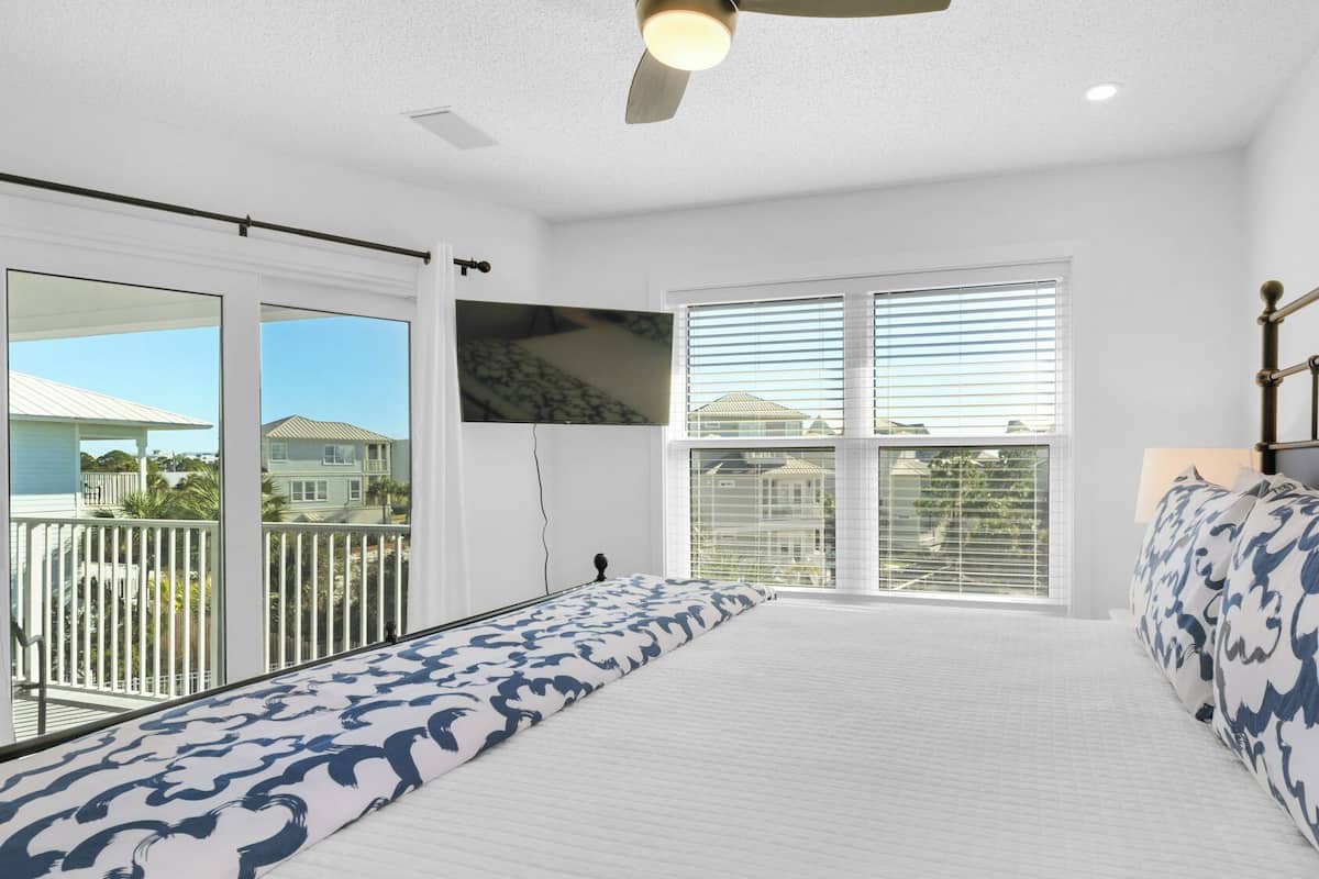 A bedroom with a large bed, a ceiling fan, a TV mounted on the wall, and a balcony with a view of houses.