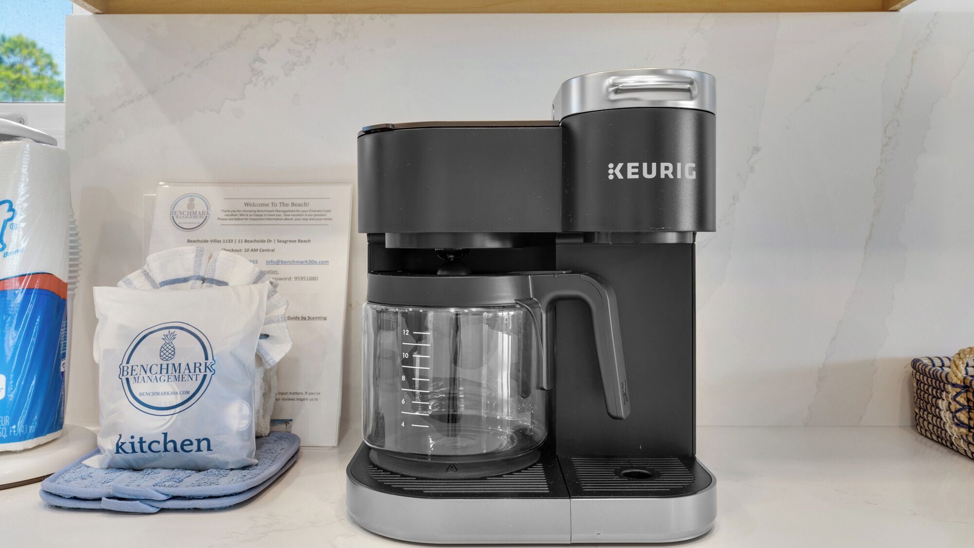 A Keurig coffee maker on a kitchen counter with a bag of Benchmark Management kitchen towels and a can of Benchmark Management coffee.
