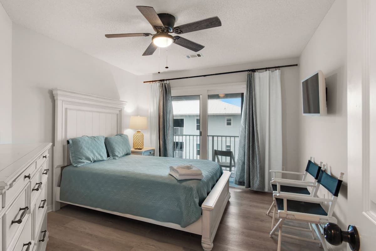 A bedroom with a bed, a nightstand, a lamp, a chair, a dresser, a ceiling fan, and a view of a balcony.