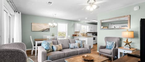 A living room with a gray sofa, a wooden coffee table, and a gray armchair. There is a kitchen in the background with white cabinets and a dining area with a white table and chairs.