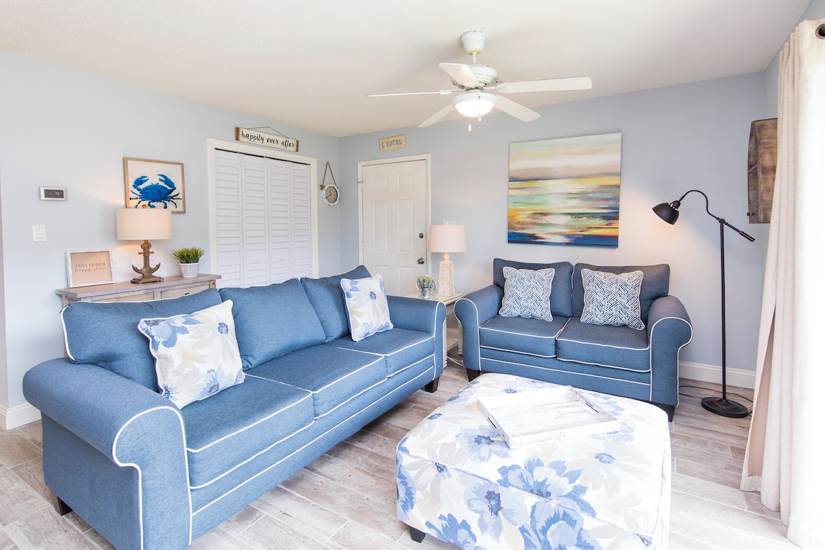A room with two blue sofas, a white ottoman, a ceiling fan, a painting, and a lamp.