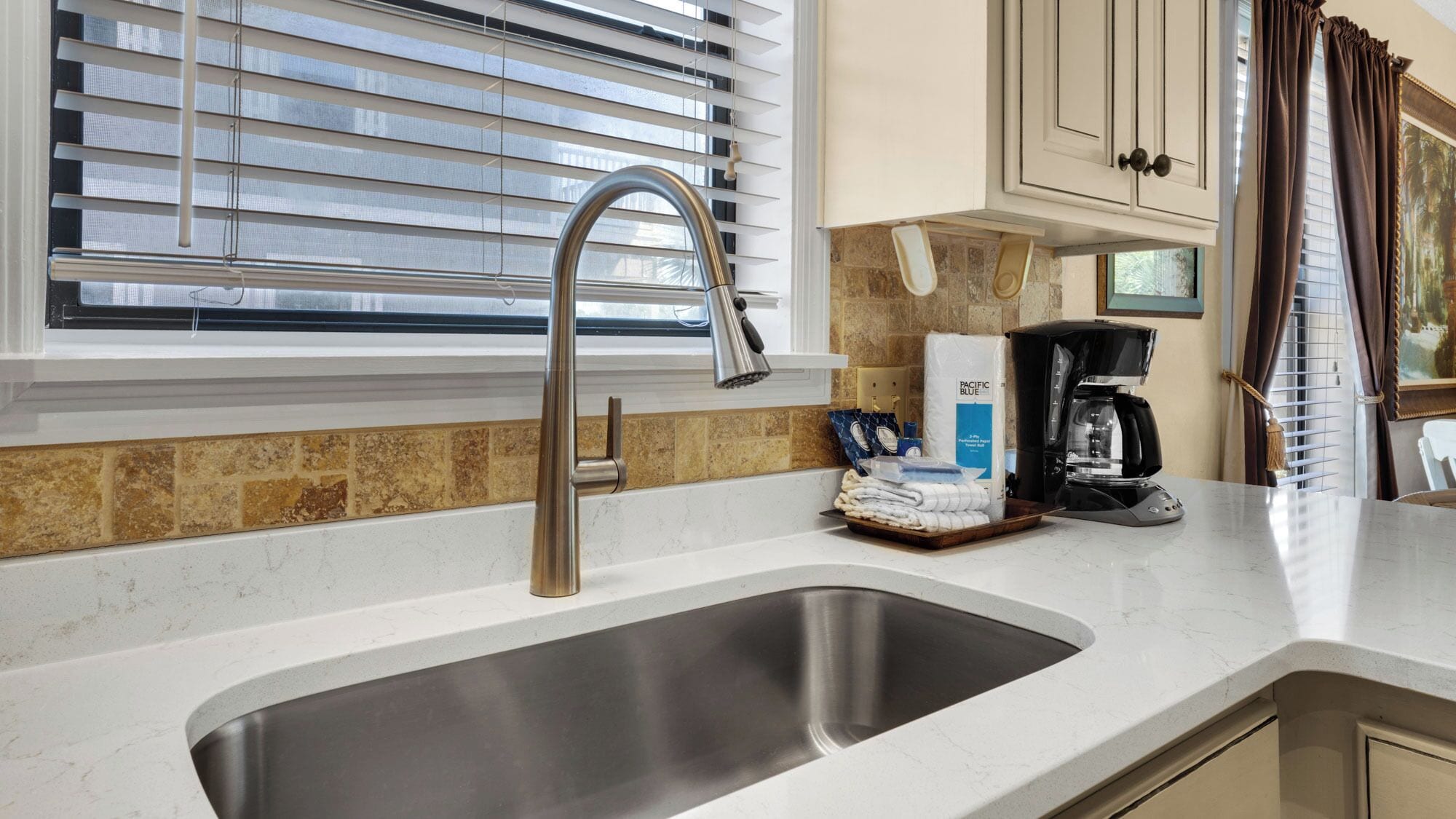 A modern kitchen with a stainless steel sink, a coffee maker, and a window with blinds.
