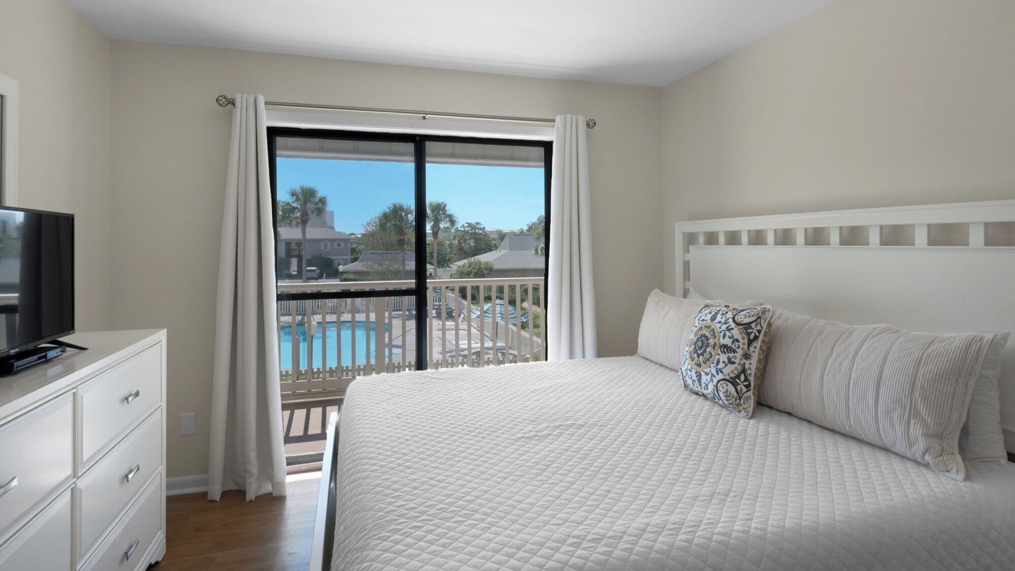 A bedroom with a bed, a TV, a dresser, and a balcony with a pool view.
