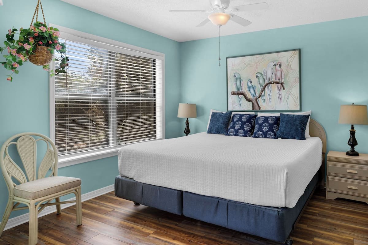 A bedroom with a bed, two bedside tables, a chair, a hanging plant, and a window with blinds.