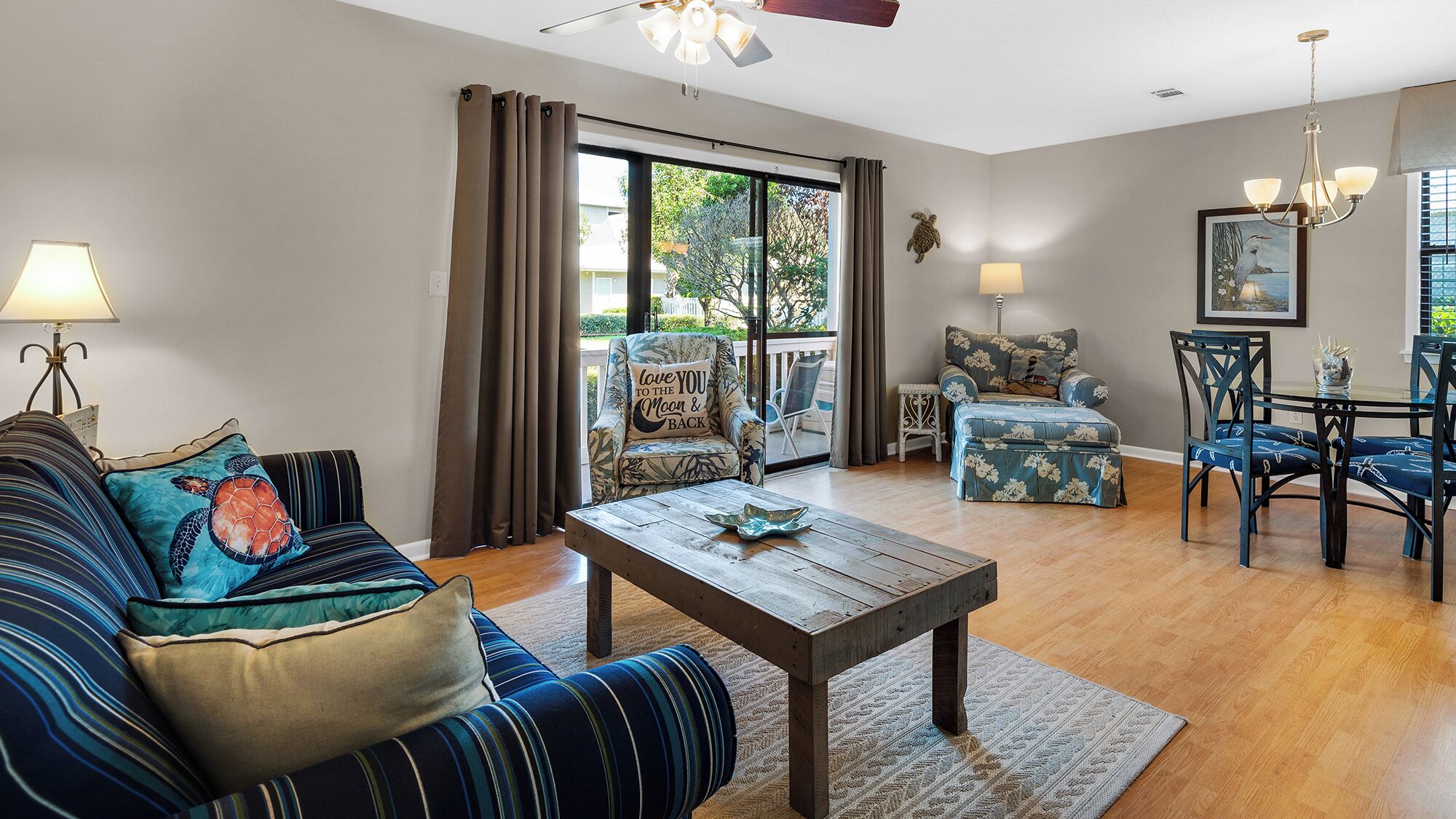 A living room with a sofa, a coffee table, a dining table with chairs, and a view through a glass door.