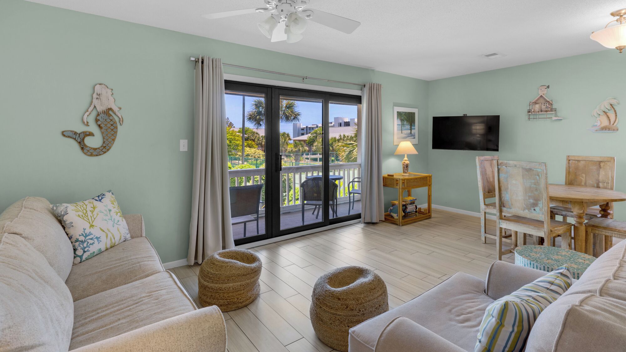 A spacious living room with a large sliding glass door leading to a balcony, a flat-screen TV mounted on the wall, and a small wooden table with a lamp.