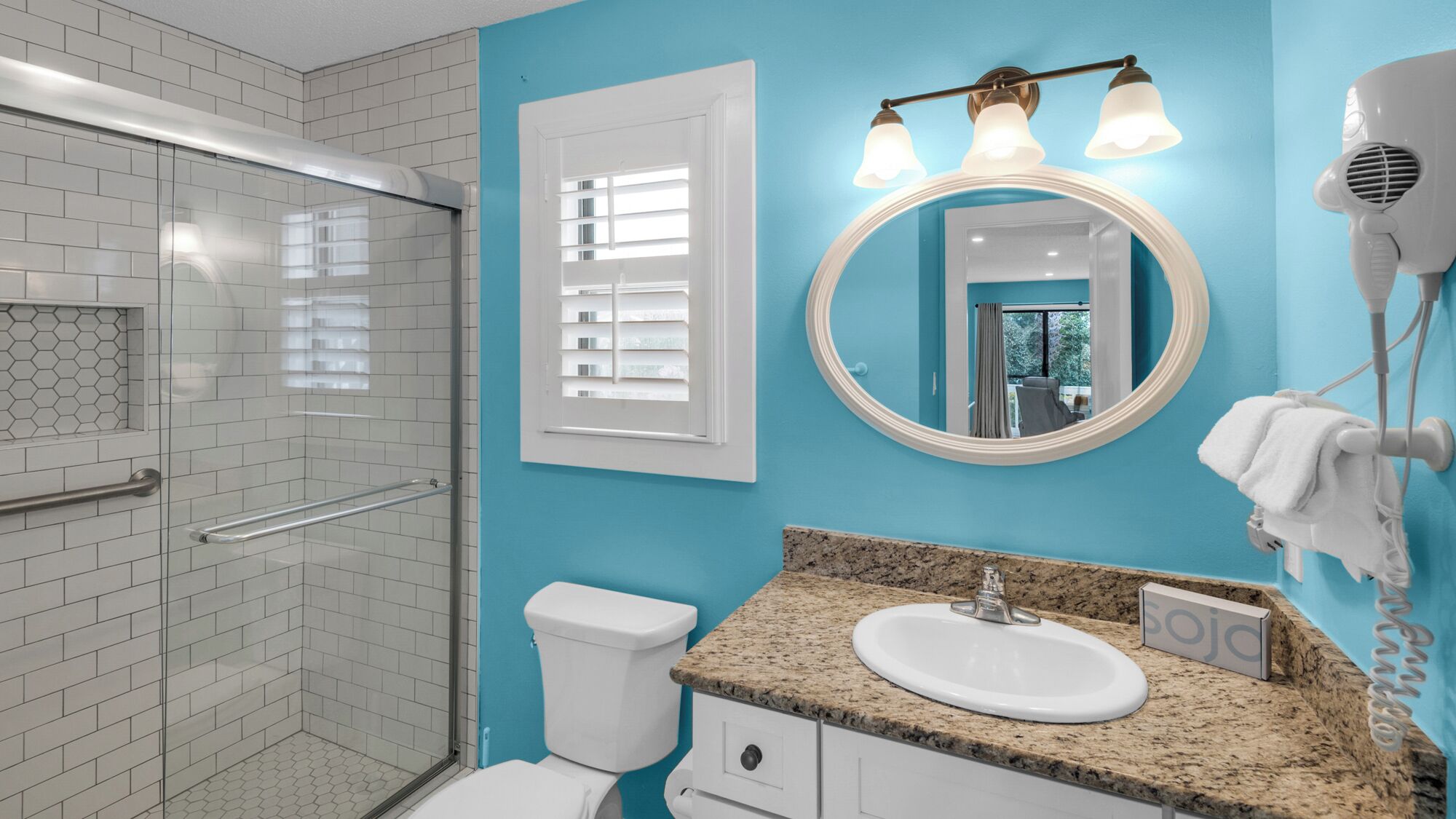 A bathroom with a glass shower enclosure, a toilet, a sink with a granite countertop, a mirror with lights, and a towel rack.