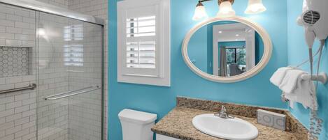 A bathroom with a glass shower enclosure, a toilet, a sink with a granite countertop, a mirror with lights, and a towel rack.