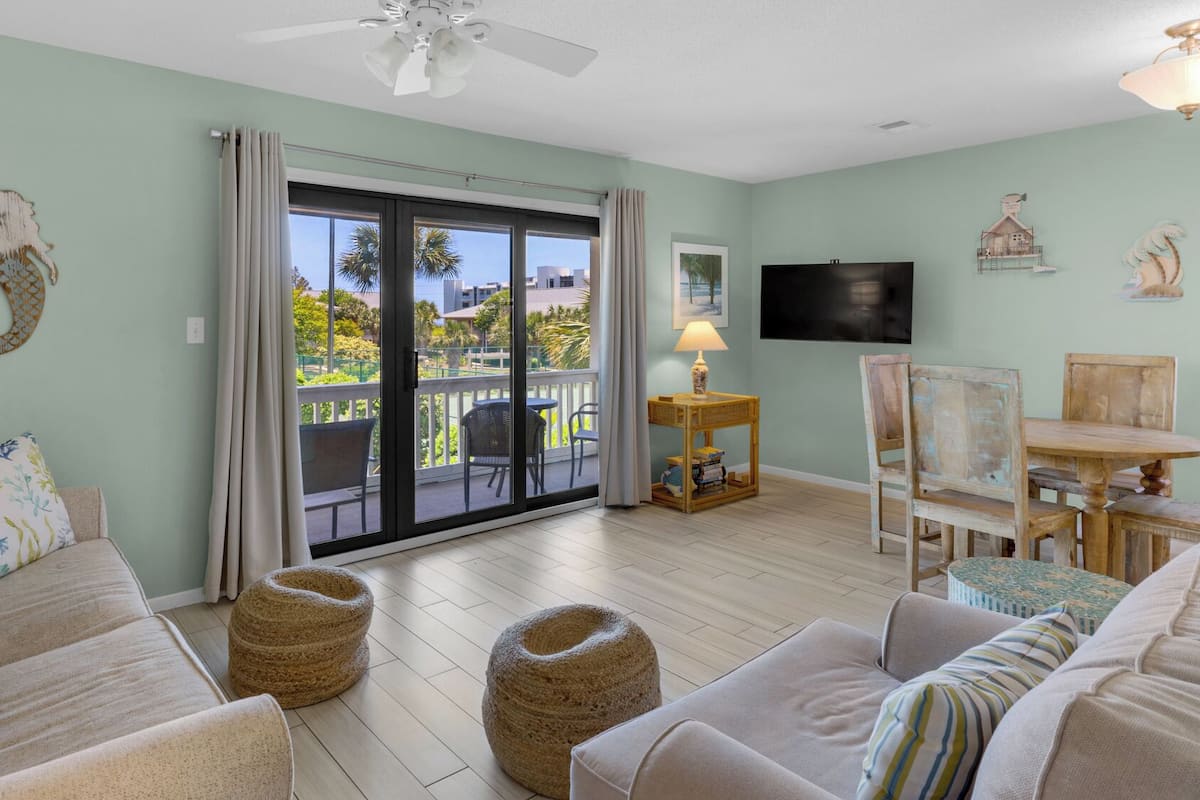 A spacious living room with a large sliding glass door leading to a balcony, a flat-screen TV mounted on the wall, and a small wooden table with a lamp.
