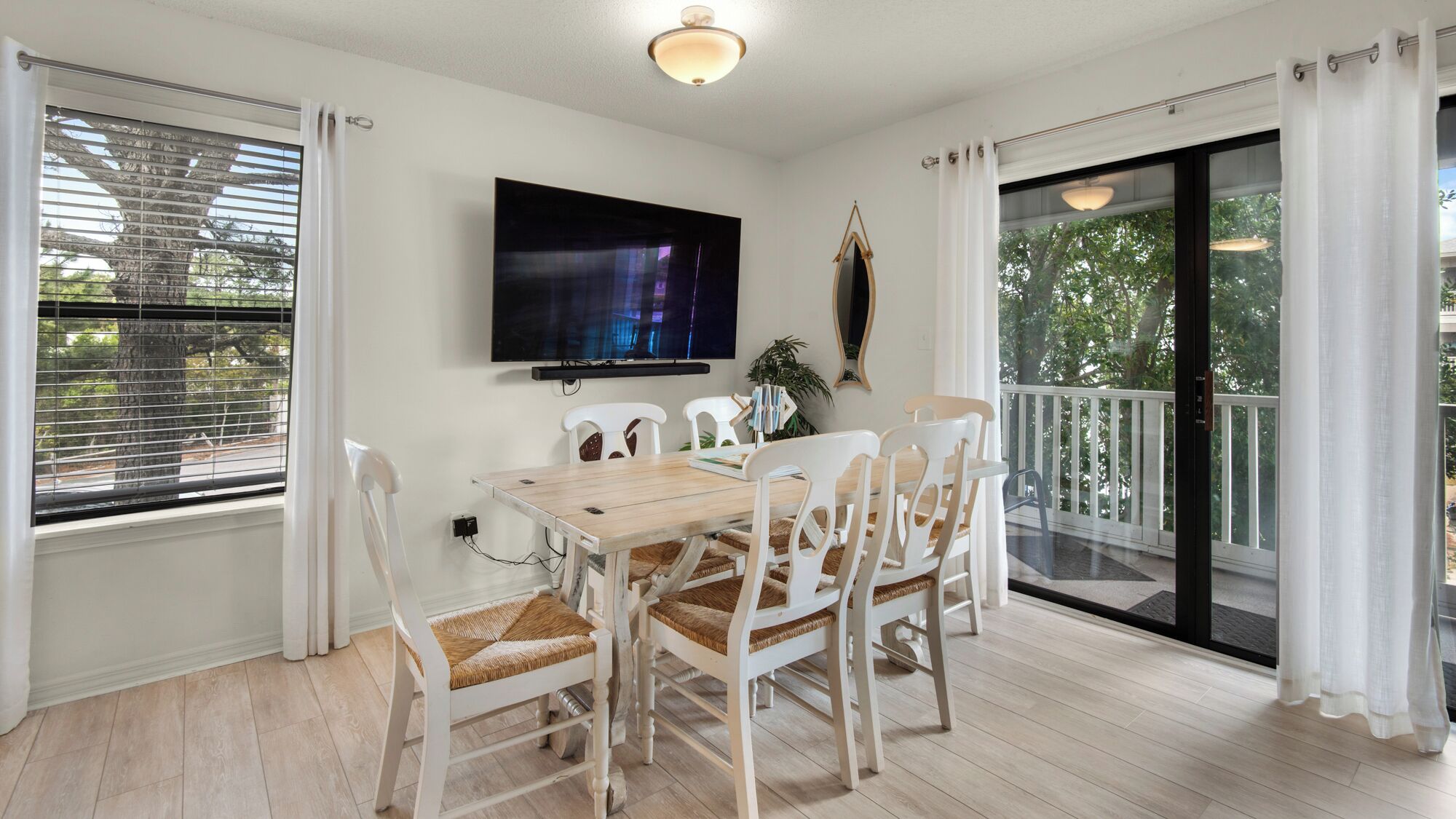 Un comedor con mesa de madera y sillas blancas, un televisor de pantalla plana en la pared, y un balcón con puerta de vidrio.