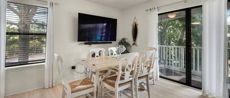 Un comedor con mesa de madera y sillas blancas, un televisor de pantalla plana en la pared, y un balcón con puerta de vidrio.