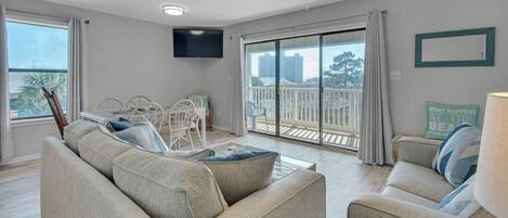 A spacious living room with a large sofa, a dining table with chairs, a flat-screen TV mounted on the wall, and a balcony with a view of the city.