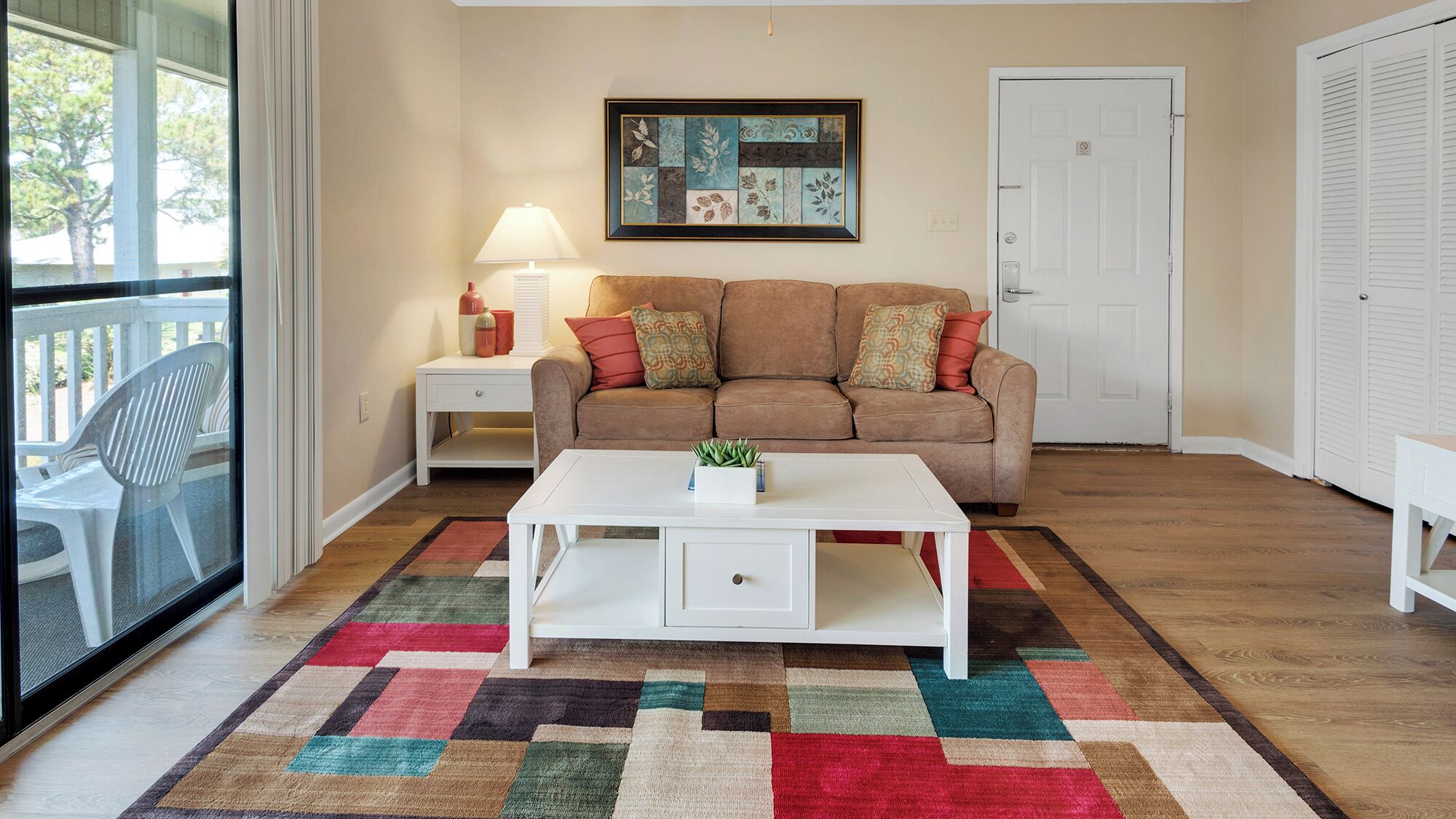 Una sala de estar con un sofá beige, una mesa de centro blanca y una alfombra colorida. Hay una obra de arte enmarcada en la pared y una puerta a la derecha.