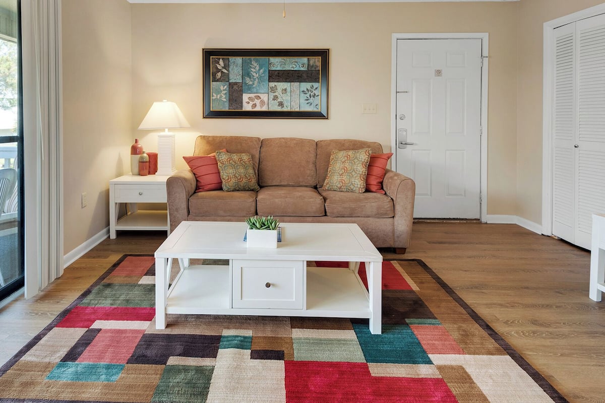 Una sala de estar con un sofá beige, una mesa de centro blanca y una alfombra colorida. Hay una obra de arte enmarcada en la pared y una puerta a la derecha.
