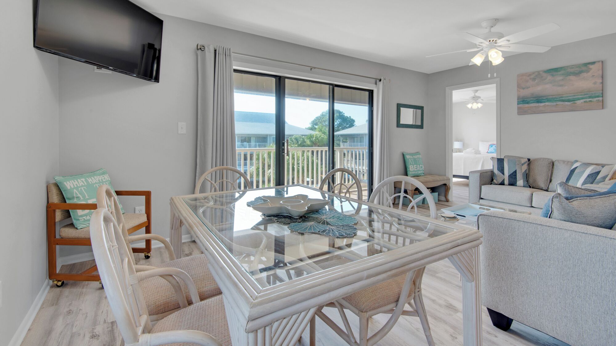 A spacious living room with a glass dining table, white wicker chairs, a grey sofa, and a ceiling fan.