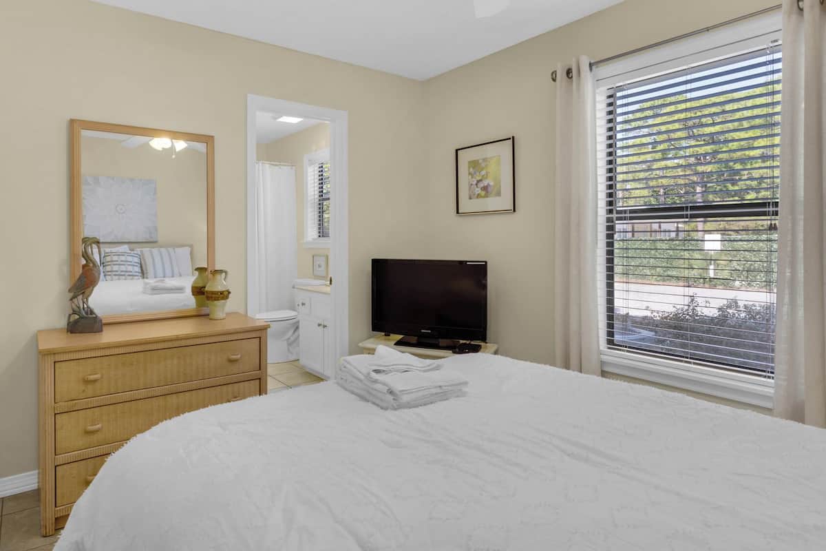 A bedroom with a bed, a dresser, a television, a window with blinds, and a bathroom visible through an open door.