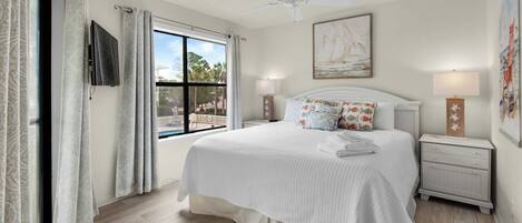 A bedroom with a large bed, a ceiling fan, a window with a view of a pool, and a painting on the wall.