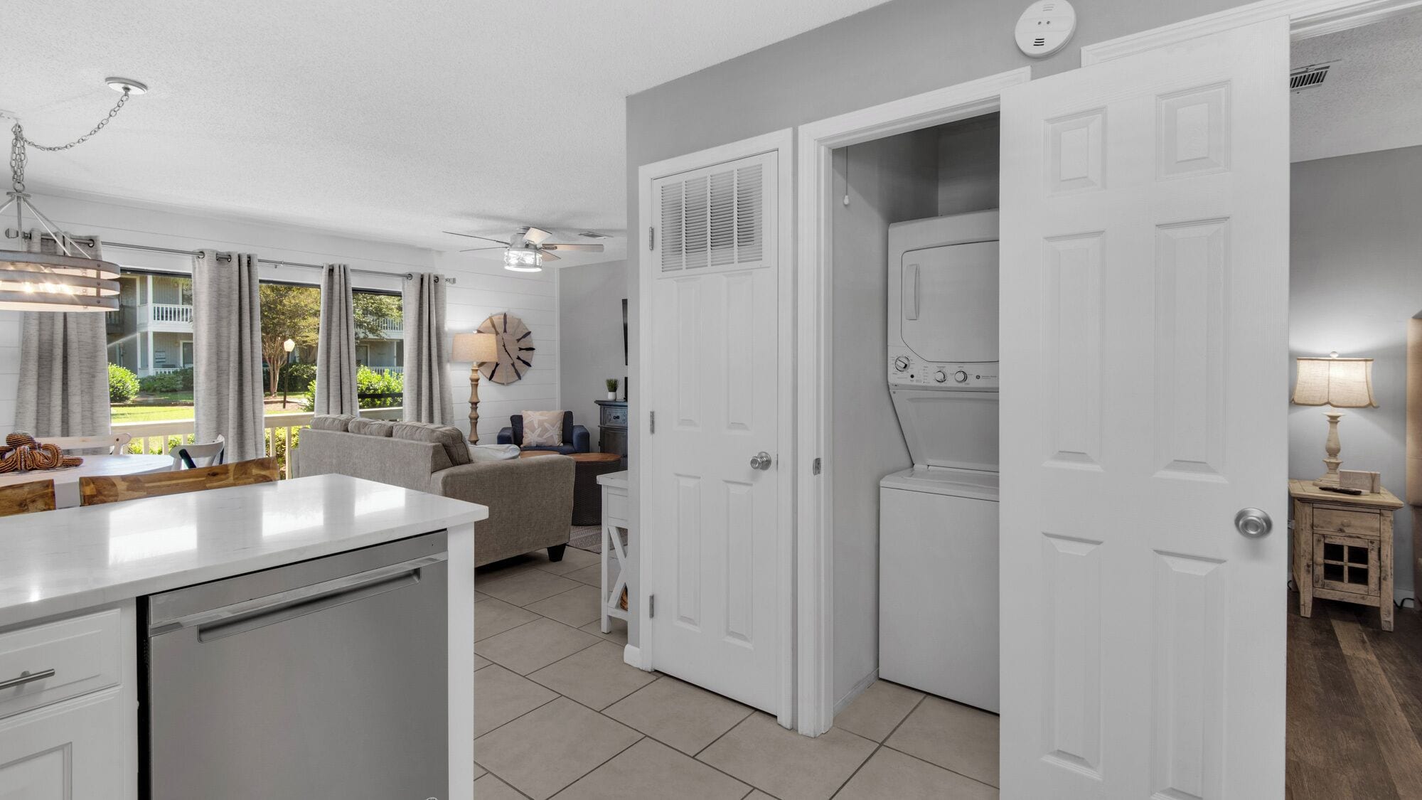 A compact kitchen with a dishwasher, a dining area with a table and chairs, a sofa, and a view of the outdoors through large windows.