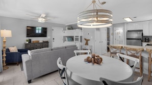A modern living room with a grey sofa, a dining table with chairs, a ceiling fan, and a kitchen area with white cabinets and appliances. - Beachwood Villas (Santa Rosa Beach)