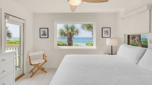 Ein Schlafzimmer mit einem großen Bett, einem Sessel, einem Nachttisch und einem Fenster mit Blick auf den Ozean und Palmen.