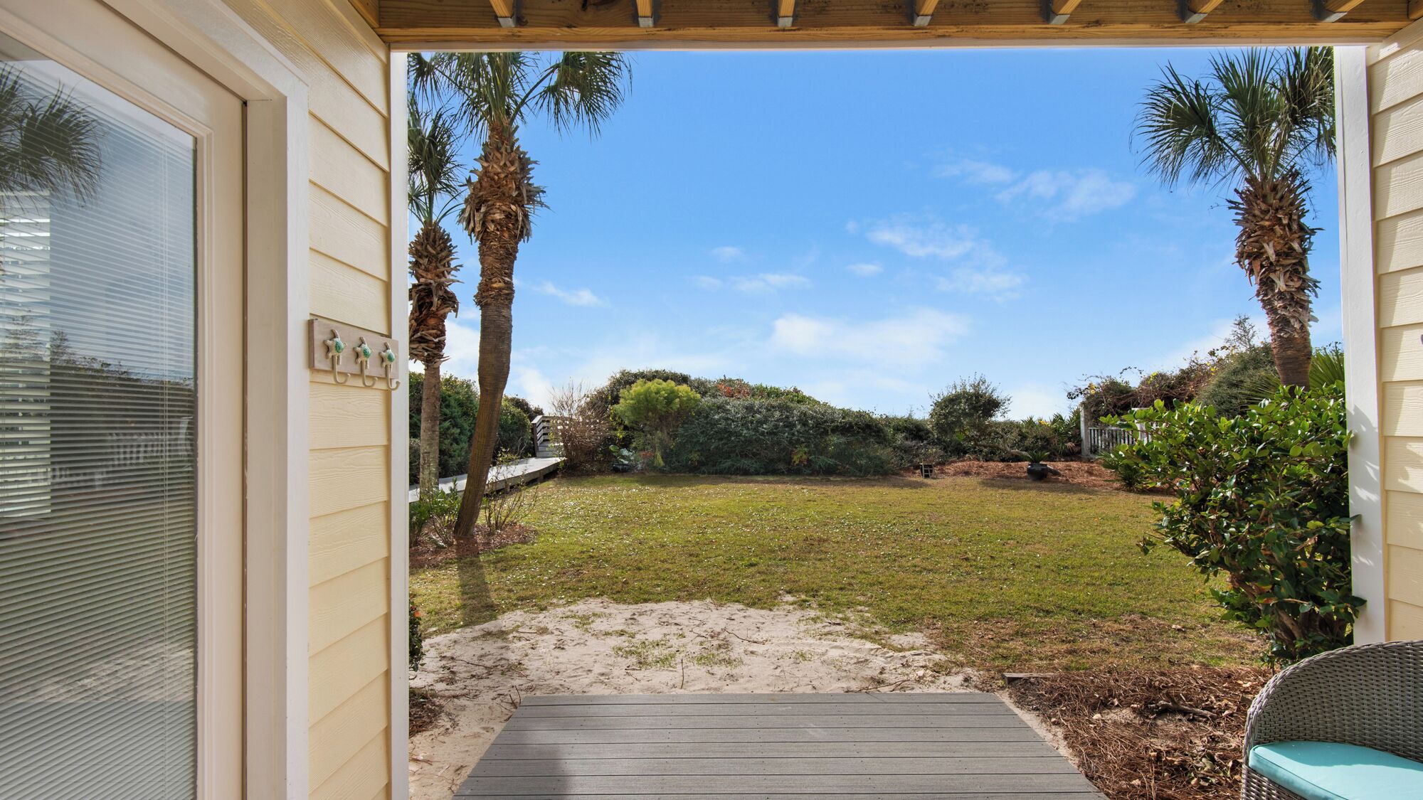 A covered patio with a wooden deck leading to a grassy yard with palm trees and a view of the sky.