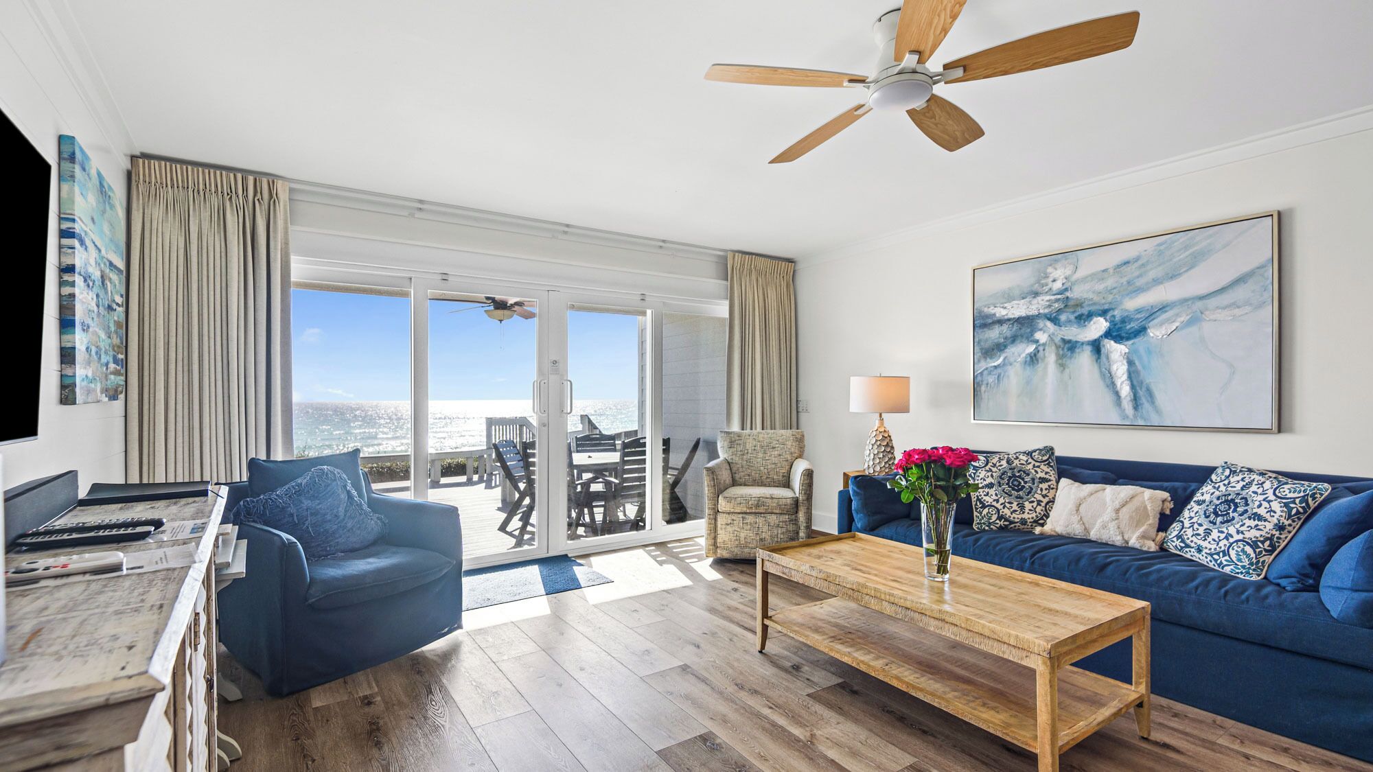 A spacious living room with a large window offering a view of the ocean, a ceiling fan, and a blue sofa with patterned cushions.