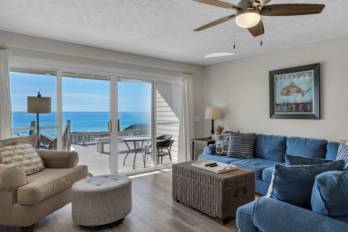 A spacious living room with a large sofa, a smaller sofa, and a round ottoman. There is a wicker basket and a table with a lamp. A ceiling fan is visible, and a framed picture hangs on the wall.