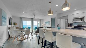 A modern kitchen with stainless steel appliances, a central island with bar stools, and a view of the ocean through large windows. - Blue Tide (Panama City Beach)