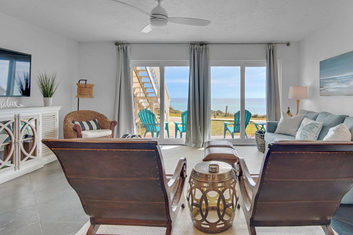 A spacious living room with a large window overlooking the ocean, a comfortable seating area with a sofa and chairs, and a dining table with chairs.