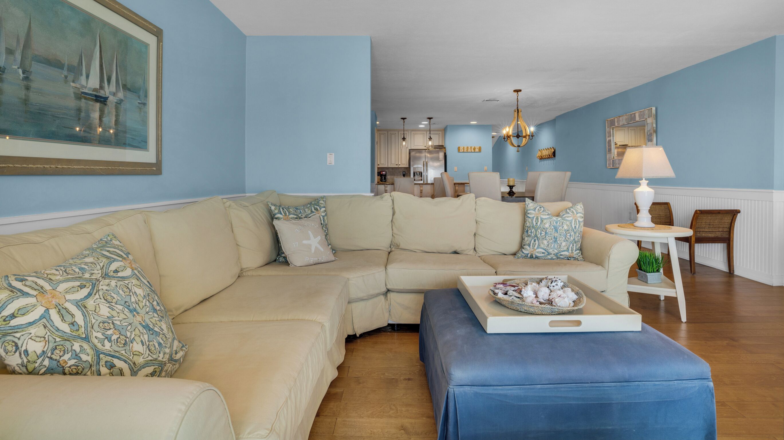 A living room with a beige sectional sofa, a blue ottoman, and a dining area with a table and chairs.