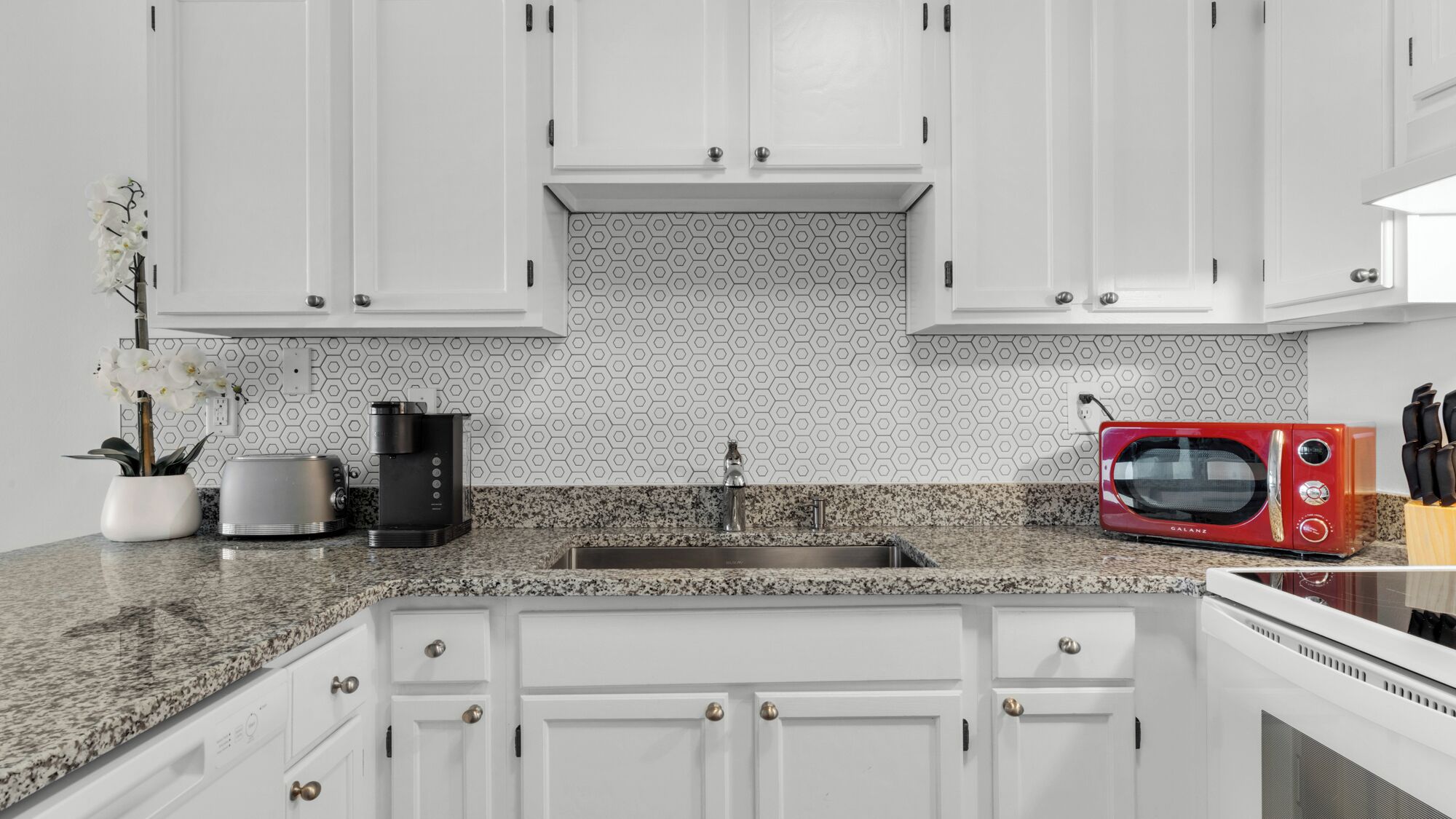 Una cocina moderna con armarios blancos, encimera de granito, cafetera, microondas rojo y un bloque para cuchillos.