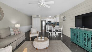 A modern living room with a sofa, a coffee table, and a TV. There is a kitchen in the background with white cabinets and a dining area with white chairs.