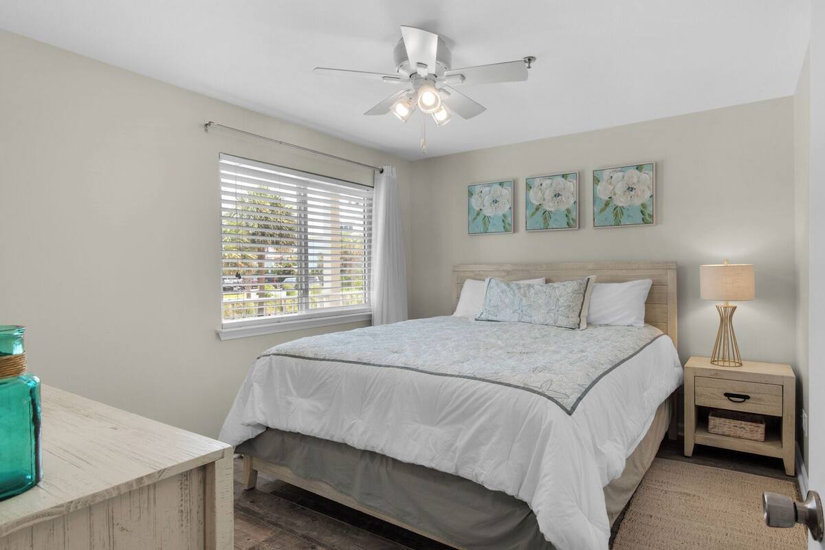 Un dormitorio con cama, mesita de noche, lámpara, ventana con persianas, ventilador de techo y decoración en la pared.