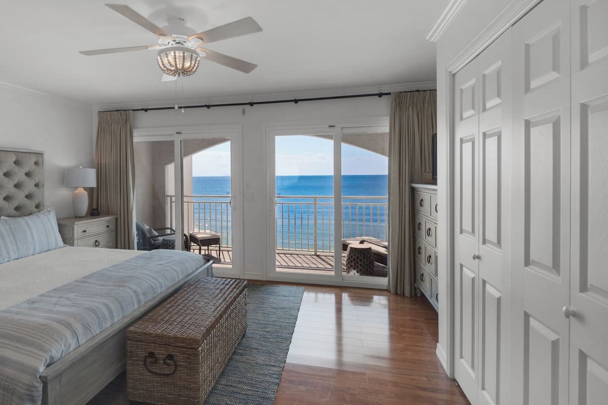Un dormitorio con una cama grande, ventilador de techo, vista al océano y balcón con mobiliario exterior.