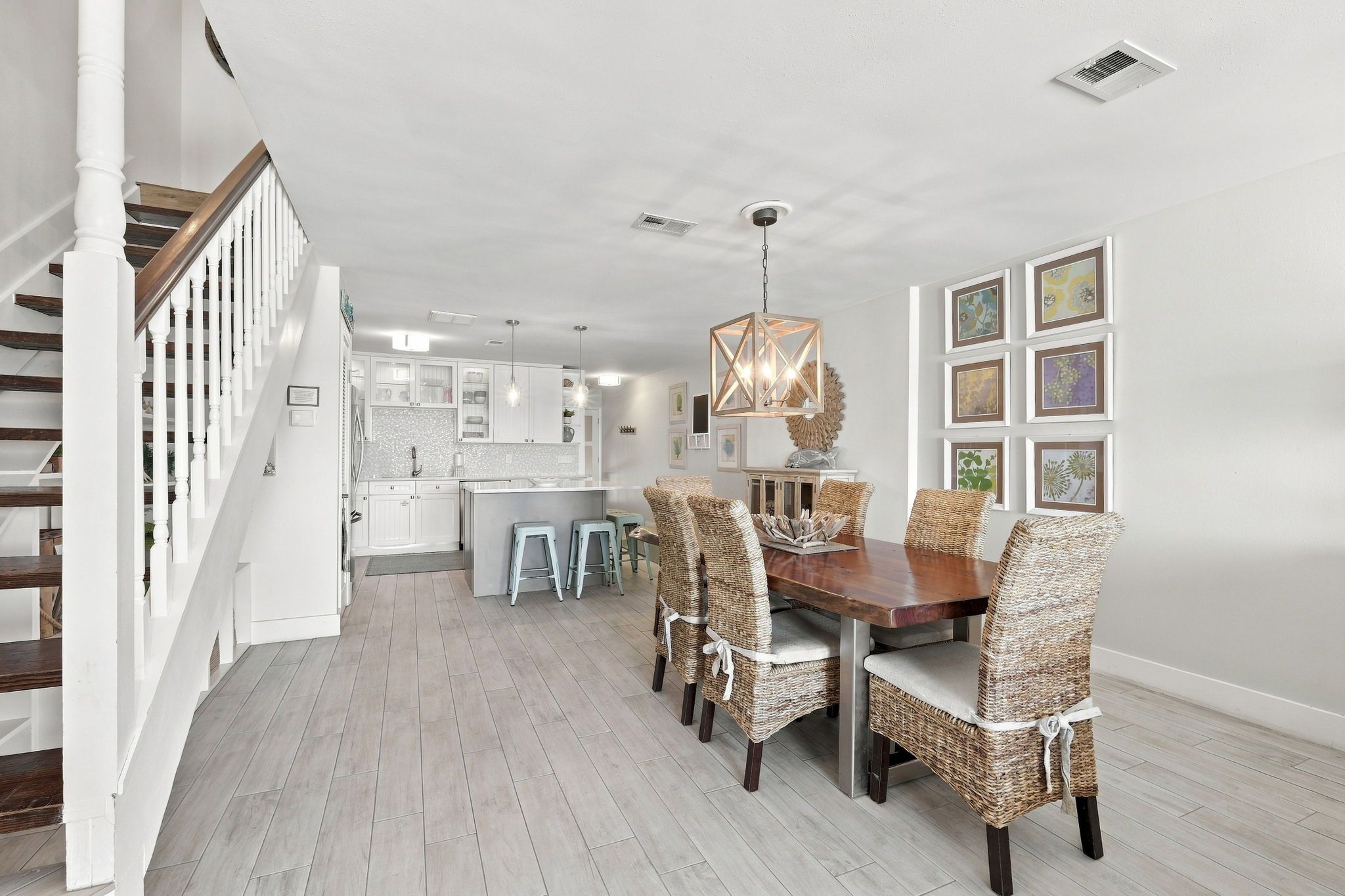 A spacious dining area with a wooden table and wicker chairs, a staircase with white balusters, and a kitchen in the background.