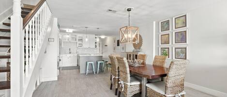 A spacious dining area with a wooden table and wicker chairs, a staircase with white balusters, and a kitchen in the background.