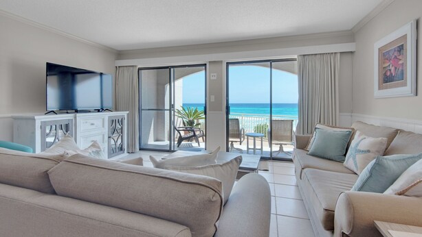 A spacious living room with a large flat-screen TV, beige sofas, and a balcony with a view of the ocean. - Dune Villas (Santa Rosa Beach)