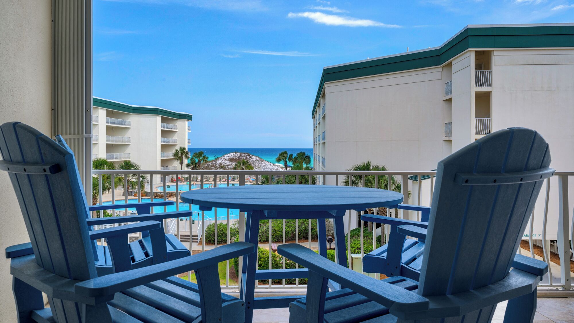 Un balcon avec des fauteuils Adirondack bleus et une table ronde, donnant sur une piscine et un bâtiment doté d’un toit végétalisé.