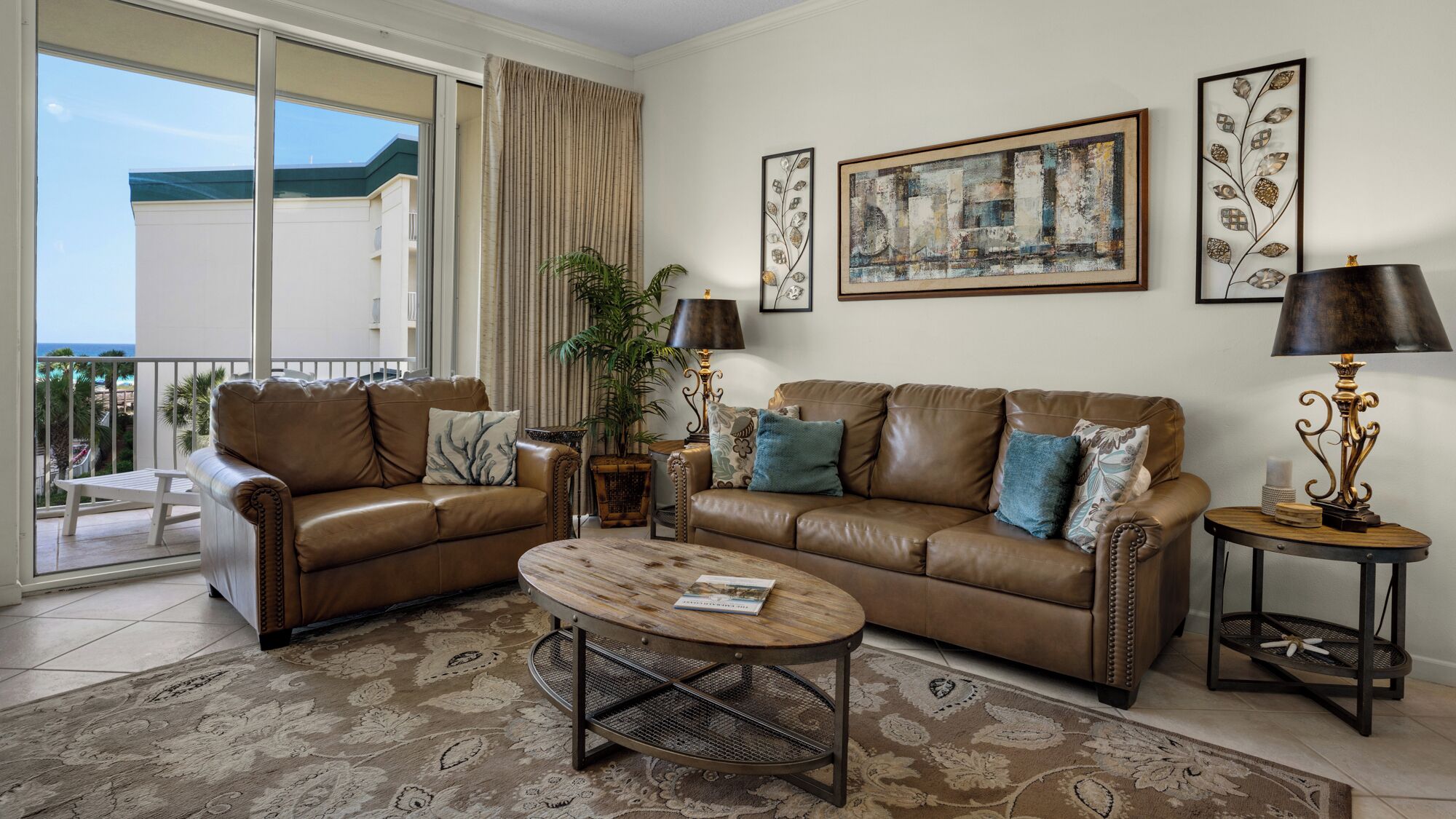 A living room with a brown leather sofa, a coffee table, and two side tables with lamps. There are two framed artworks on the wall and a large window with a view of a building and a balcony.