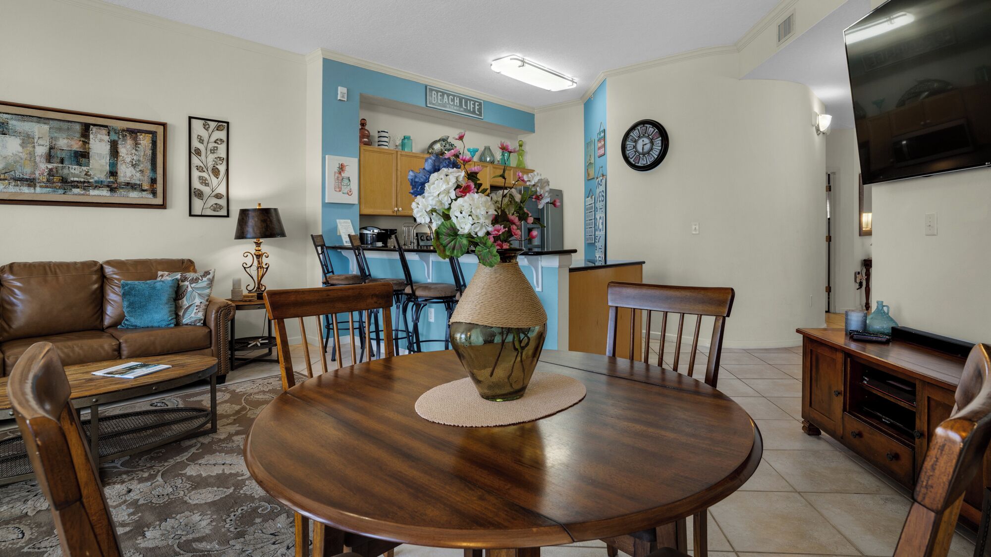 A living room with a brown leather sofa, a wooden dining table with chairs, a vase with flowers, and a kitchen area in the background.