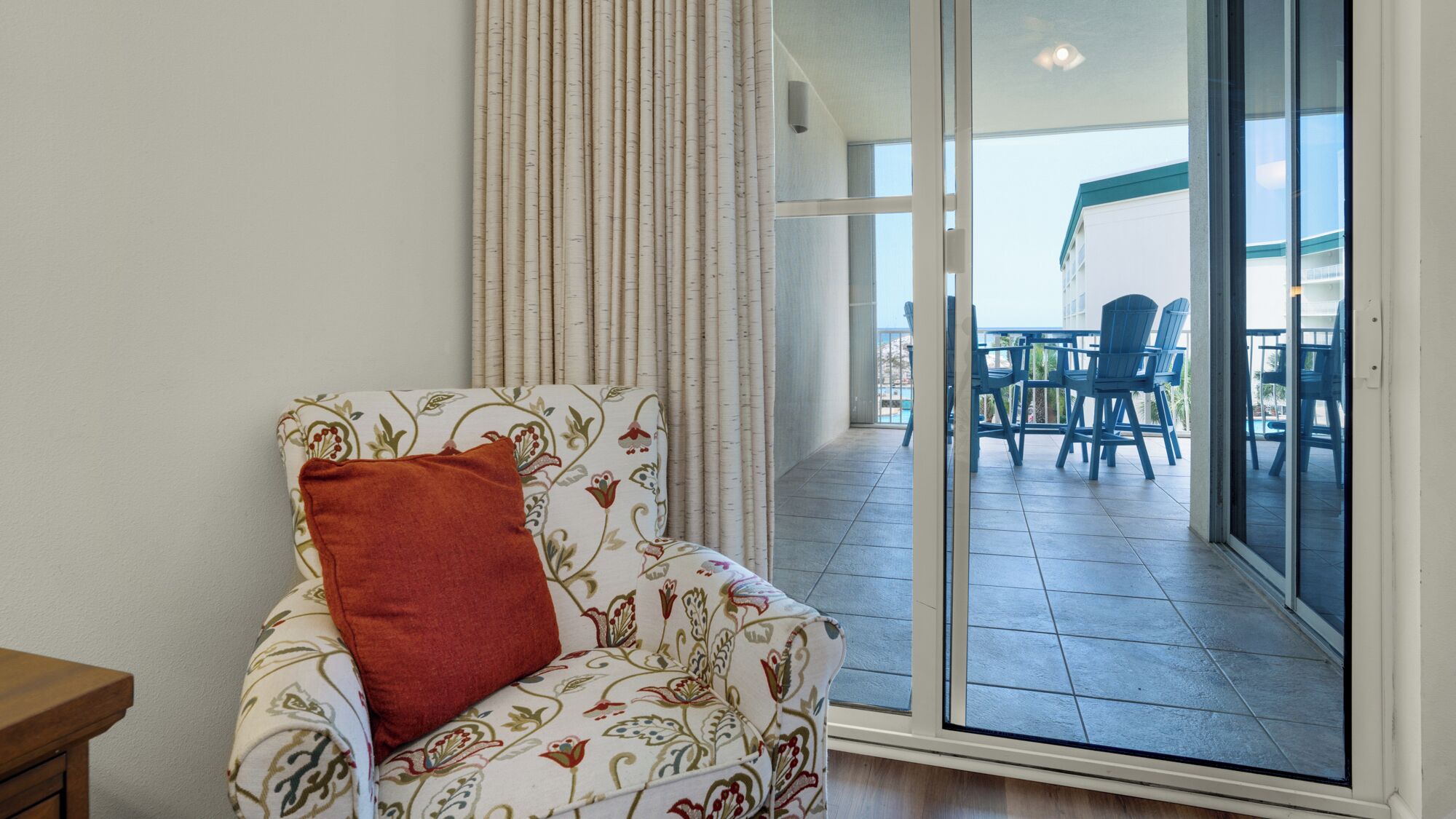 A room with a sofa and a red pillow, a sliding glass door leading to an outdoor area with blue chairs and a table.